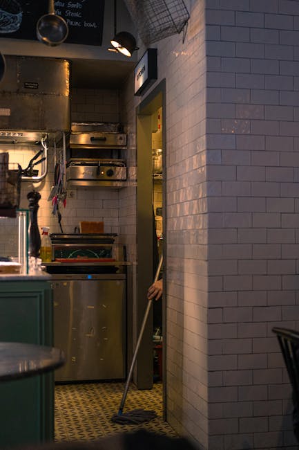 Interior view of a commercial kitchen with white tiled walls and vintage yellow-patterned flooring. A person, partially visible, is mopping the floor near a doorway. Visible surfaces include a stainle