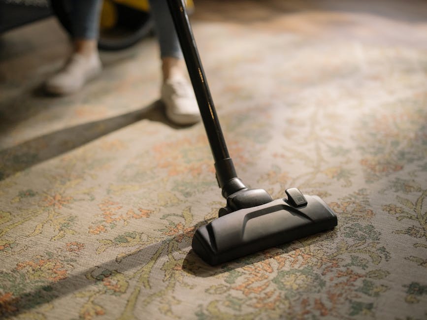 A professional cleaner dressed in a white protective suit, face mask, and blue gloves is engaged in deep cleaning a beige, high-pile carpet in a well-lit, modern residential living room. The cleaner i