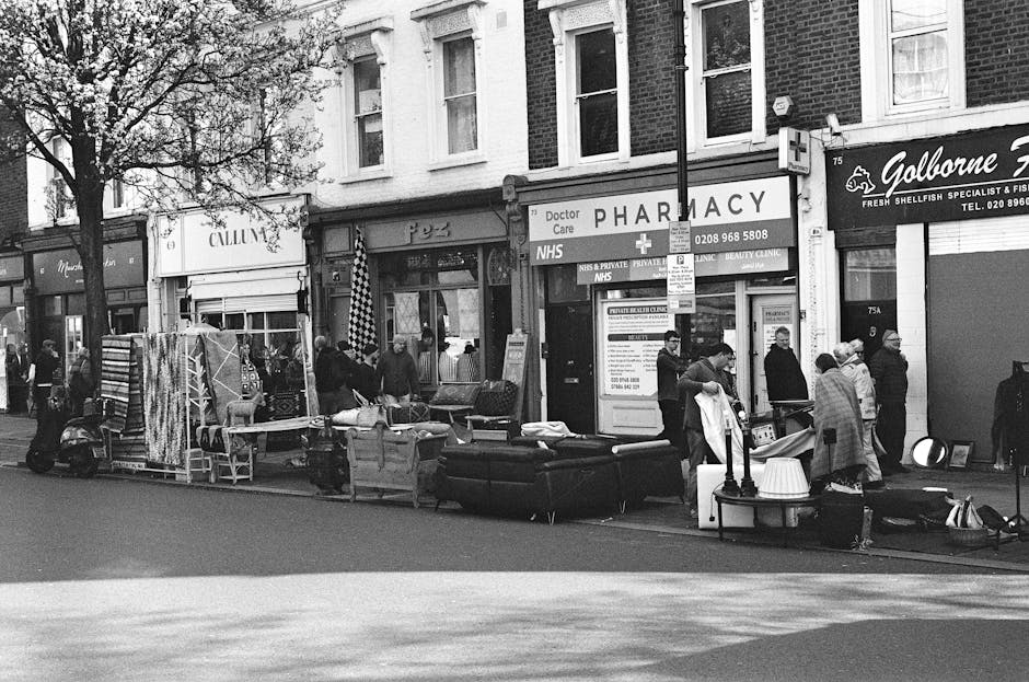 A black-and-white street scene in Clerkenwell showing storefronts and pedestrians. The image includes a pharmacy with a sign reading 'Doctor Care' and 'NHS,' along with other shops such as a fresh sea